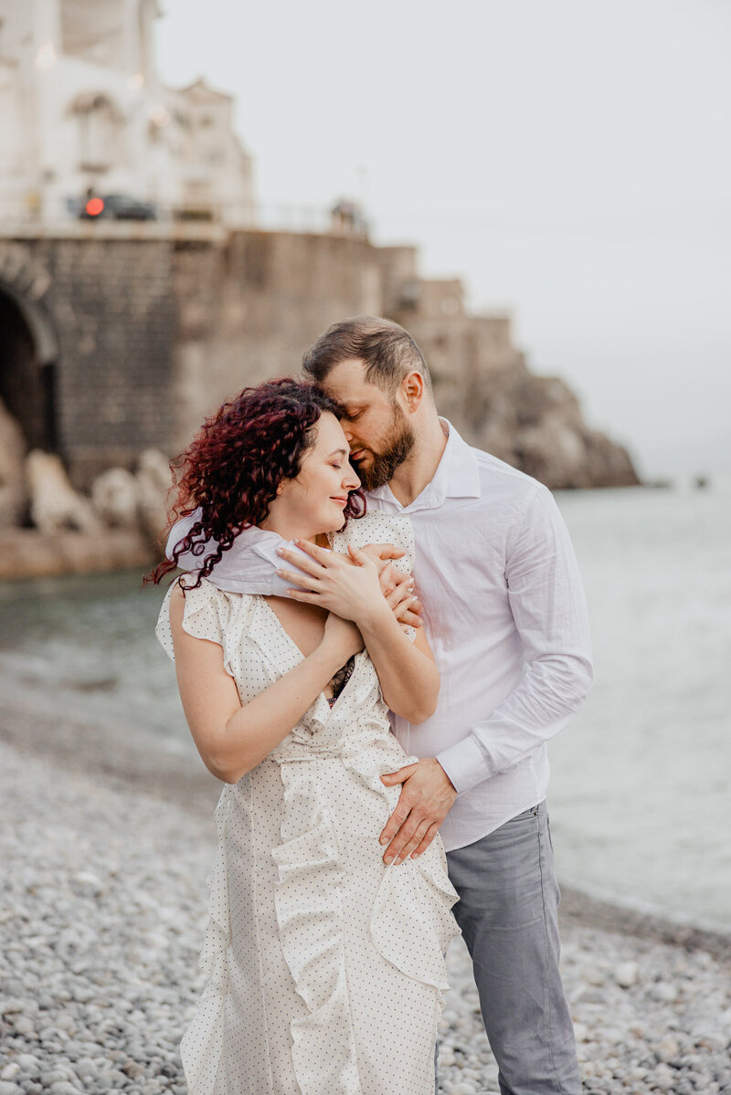 A couple embracing lovingly on a rocky beach with a historic stone bridge in the background, photographed by a UK wedding photographer and videographer.