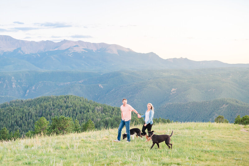 Couple walking with two black dogs on a green hill overlooking Pikes Peak.