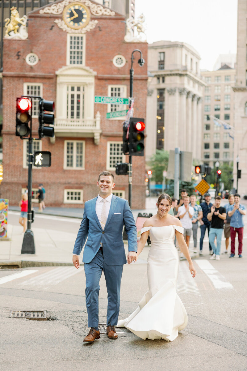 Married couple walking along a crosswalk as people cheer them on from behind.