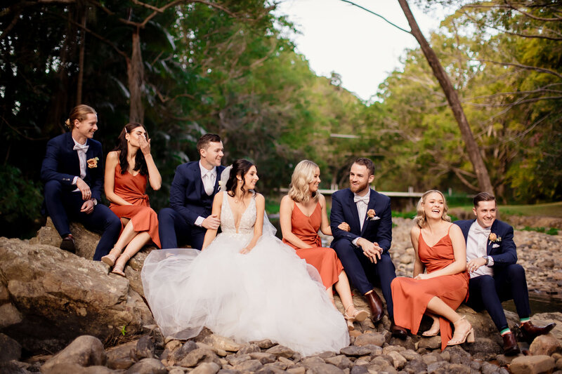 Wedding couple, bridesmaids and groomsmen, are sitting on a rock alongside a river