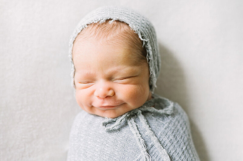 A smiling newborn baby in a gray swaddle with a matching gray bonnet.