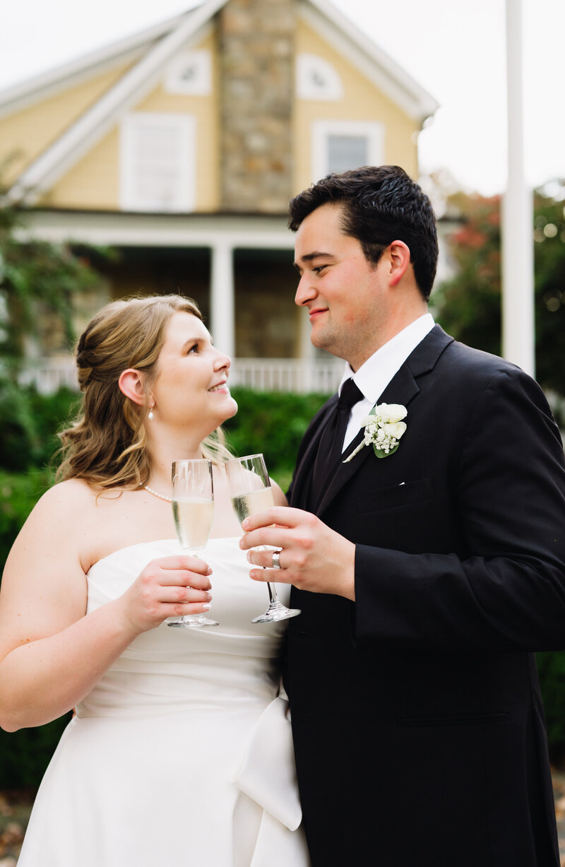 bride and groom both holding a glass of champagne and toasting as they smile to one another outside of their shenandoah wedding venue captured by charlottesville wedding photographers