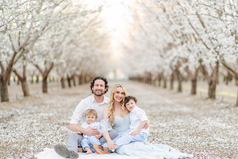 A family of four smiles in a field of almond blossoms.
