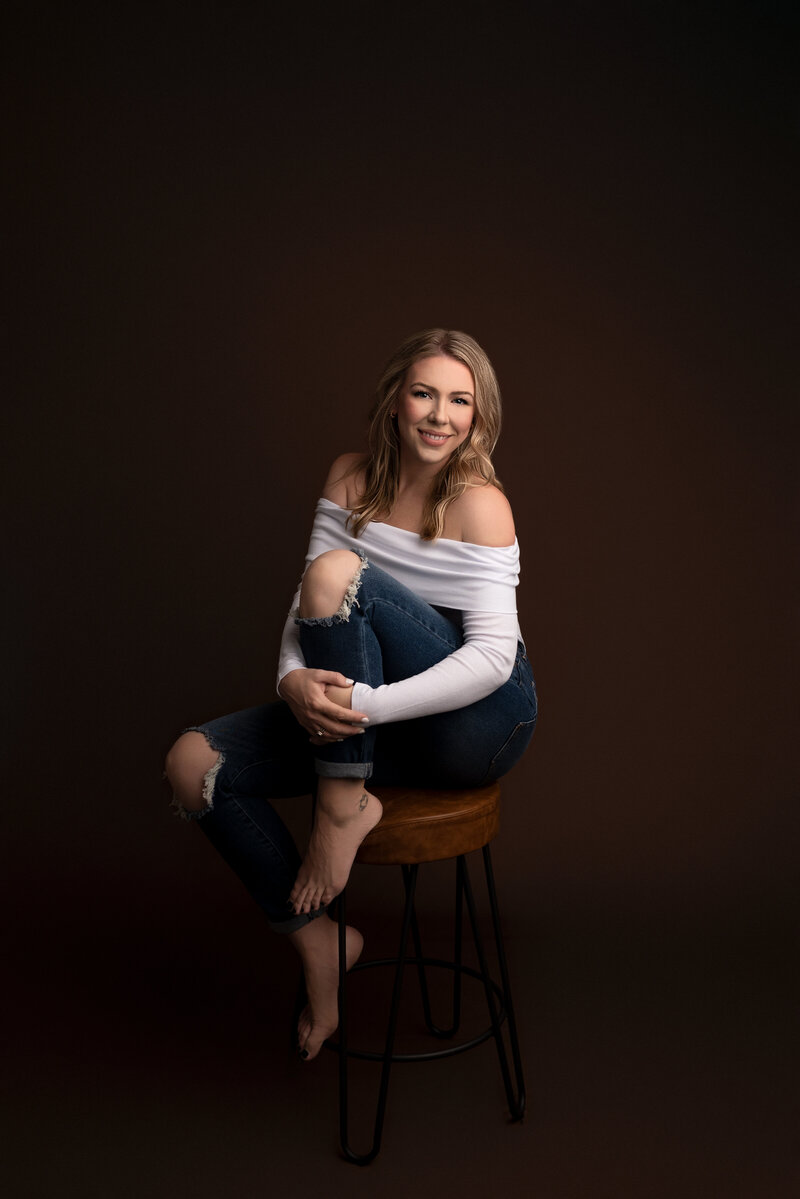 Katie Marhsall, New Jersey's best maternity and newborn photographer poses for a studio headshot. She is sitting on a still with her foreleg resting atop of the seat and hugging her fore leg. She is smiling at the camera.