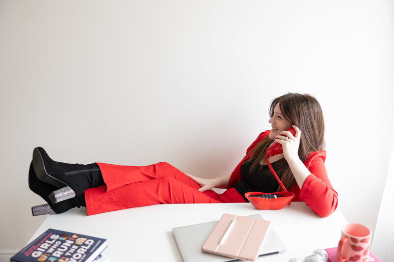 Emily in a red suit holding a red lip phone up to her ear with her feet on a white desk. There is an ipad, lap top and books on the desk.