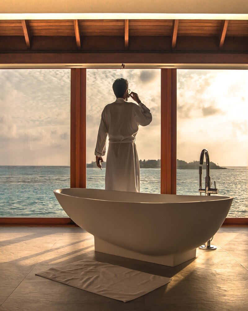 A man stands in front of large windows with a free standing bathtub behind him. It's sunset and he's standing overlooking the water in a robe.