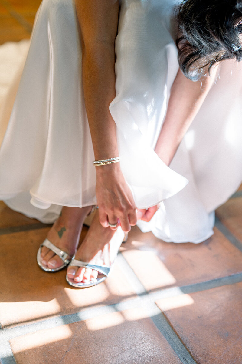 Close-up of bride slipping into strappy wedding heels in warm terracotta-tiled room filled with morning light