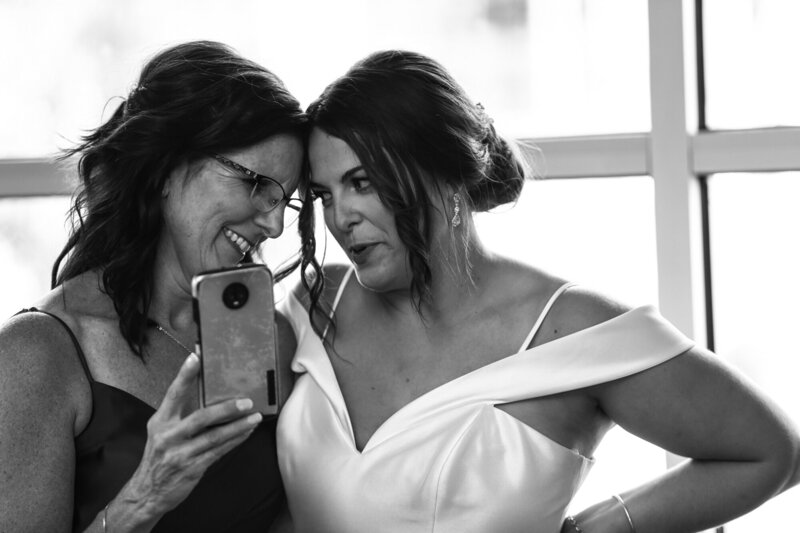 Bride and her mother stand smiling with foreheads pressed together  and reading a message on a cell phone