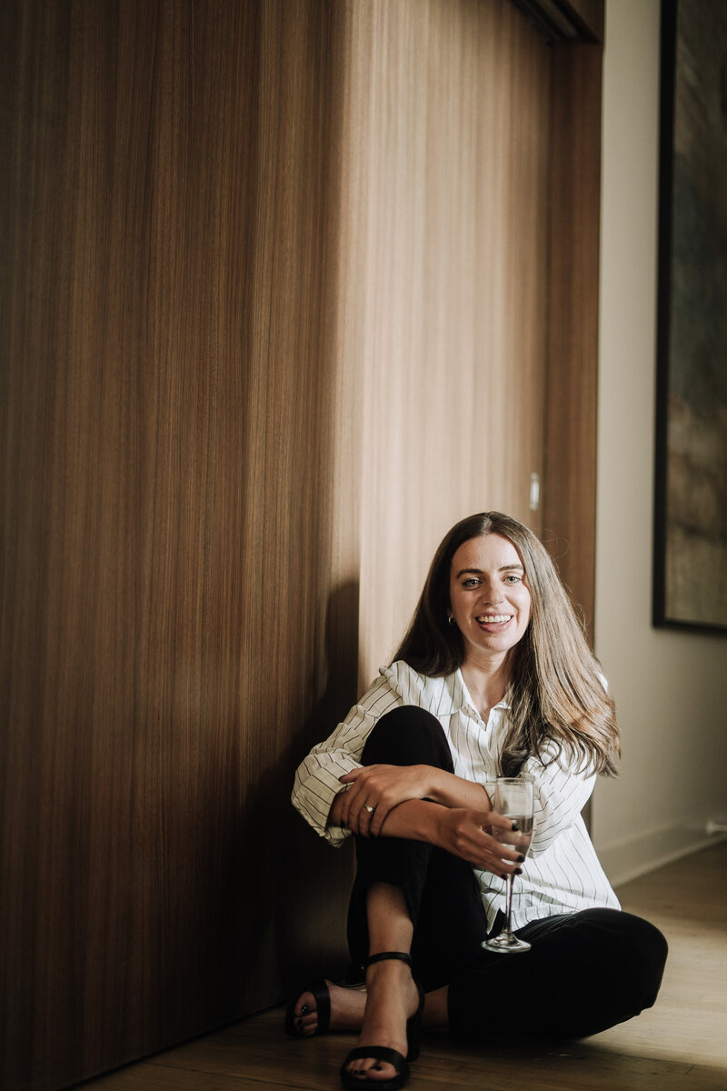 Portrait of creative director Amanda Burg wearing white button-up top, black pants, and black heels, holding a glass of champagne with warm natural sunlight on her face
