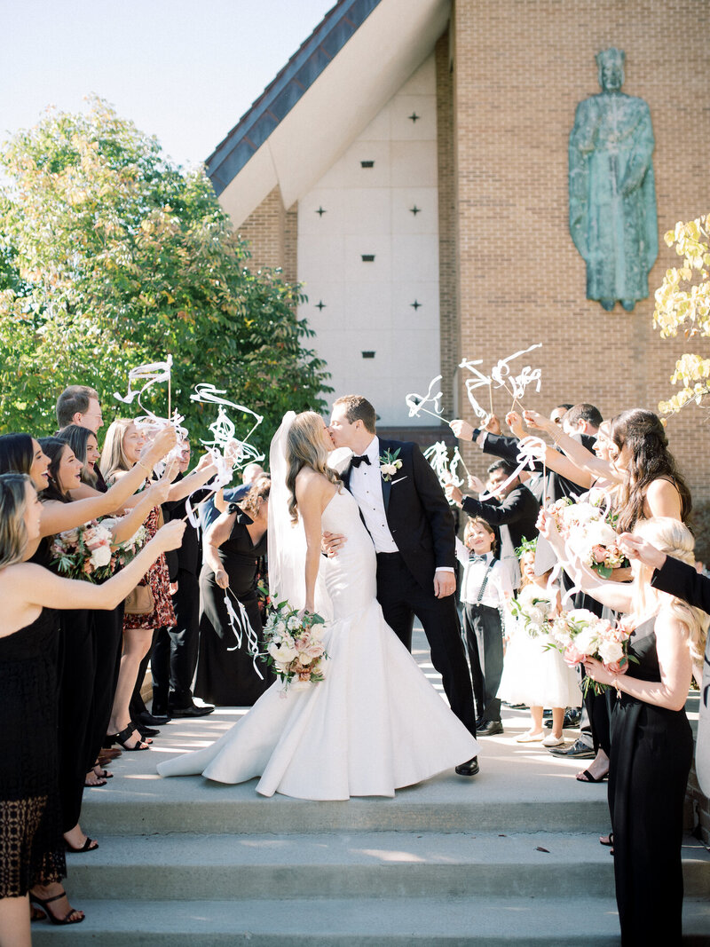 A bride and groom kissing as they leave the church and their guests wave ribbons around them