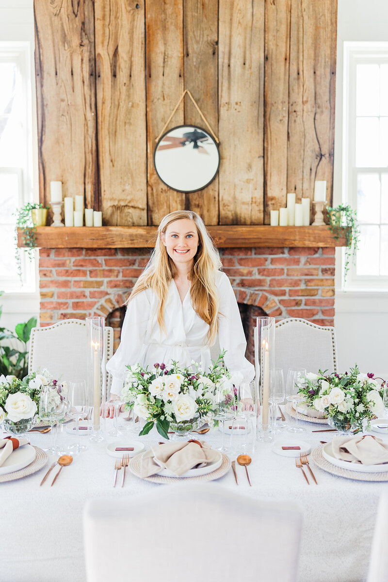 Washinton DC Day of Wedding Coordinator Blue Sapphire Events poses in front of a rustic fireplace and white wedding tablescape featuring white floral, linen napkins, tall candles, and gold flatware.