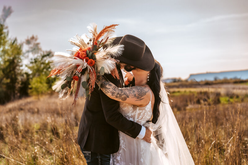 Family Photographer,  a man and woman kiss in a dry grassy field, she is in her wedding dress, he wears a cowboy hat