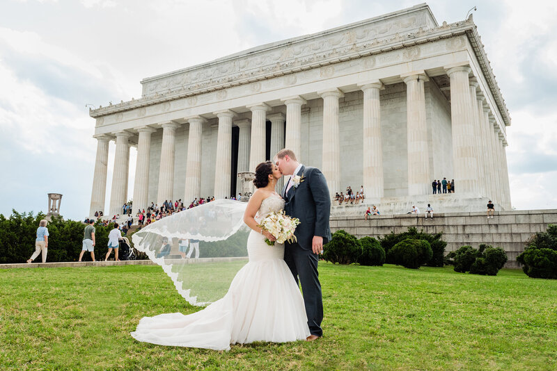 Dramatic photo of a bride and groom kissing in front of the Lincoln Memorial with her veil blowing in the wind on their wedding day. Captured by a DC wedding photographer