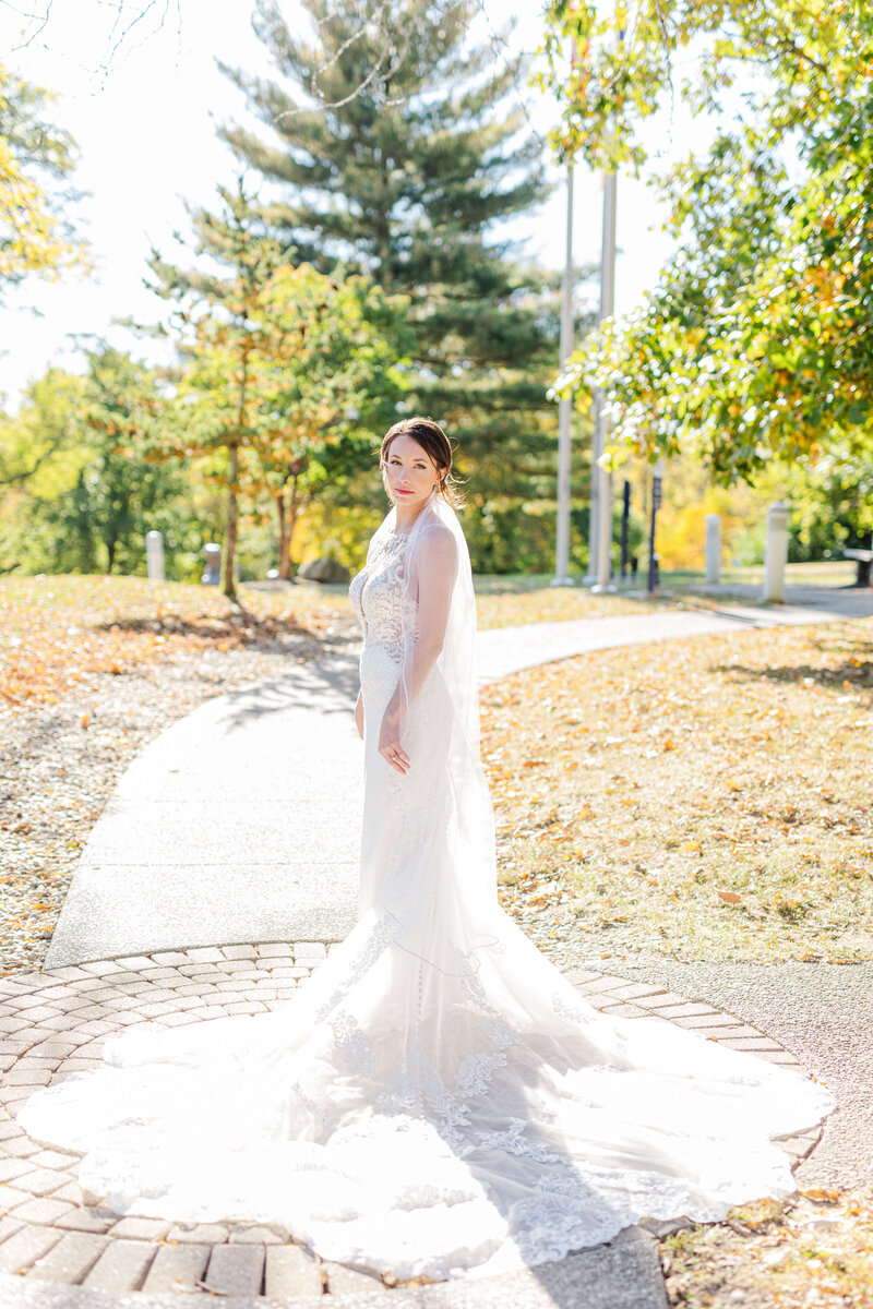 A bride is standing in a park as the sun glows through her dress. It is a beautfiul sunny day and it's fall, so the trees are yellow.