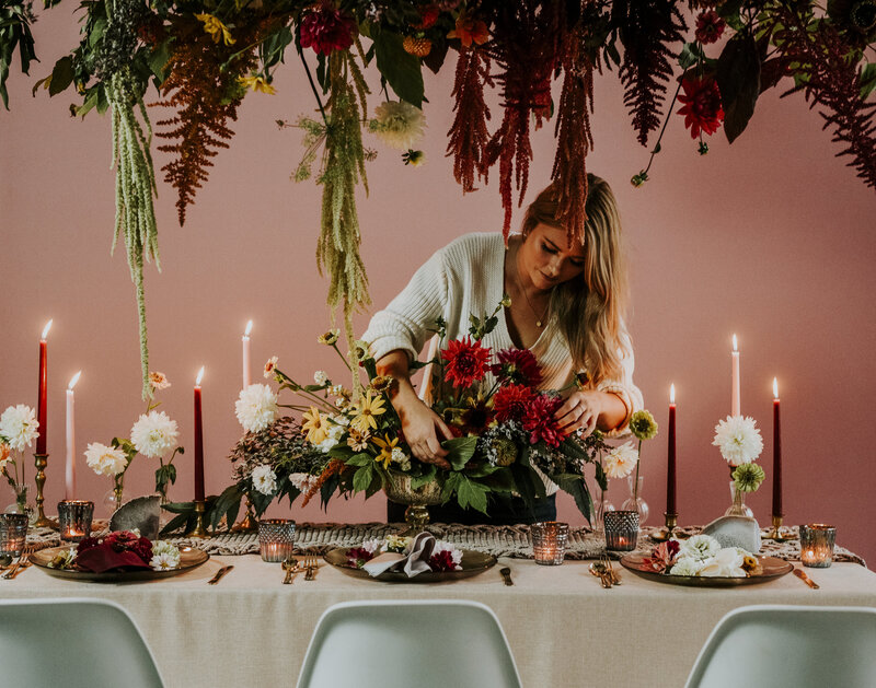 Brittany Frid lights a candle on a 70's inspired wedding guest table featuring macrame table runners, burgundy tapered candles, earthly local flowers , a massive floral ceiling installation hanging over the table and a muted pink backdrop.