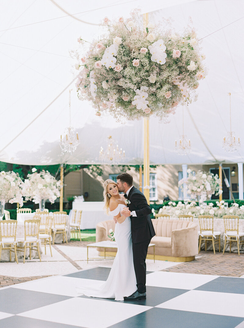 Groom kisses brides cheek on black and white checkerboard dance floor under hanging pink and white floral installation. Charleston outdoor fall wedding reception.