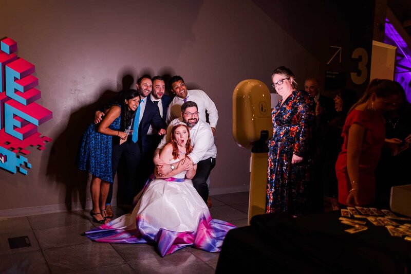 Bride and groom, surrounded by wedding party, taking a photo in front a photo booth, and their faces are illuminated by its light