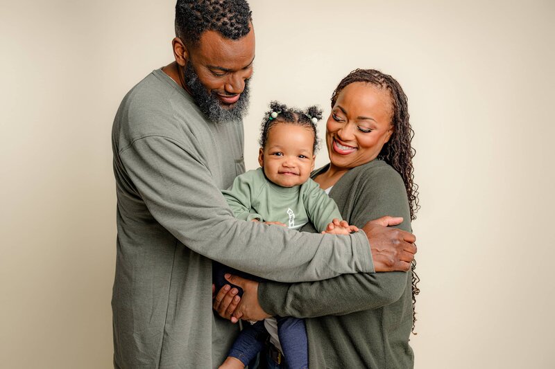 family of 3 cuddled together  dressed i greens as they look down at their one year old daughter