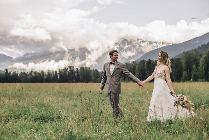 Bride and Groom holding hands walking through a field next to a mountain