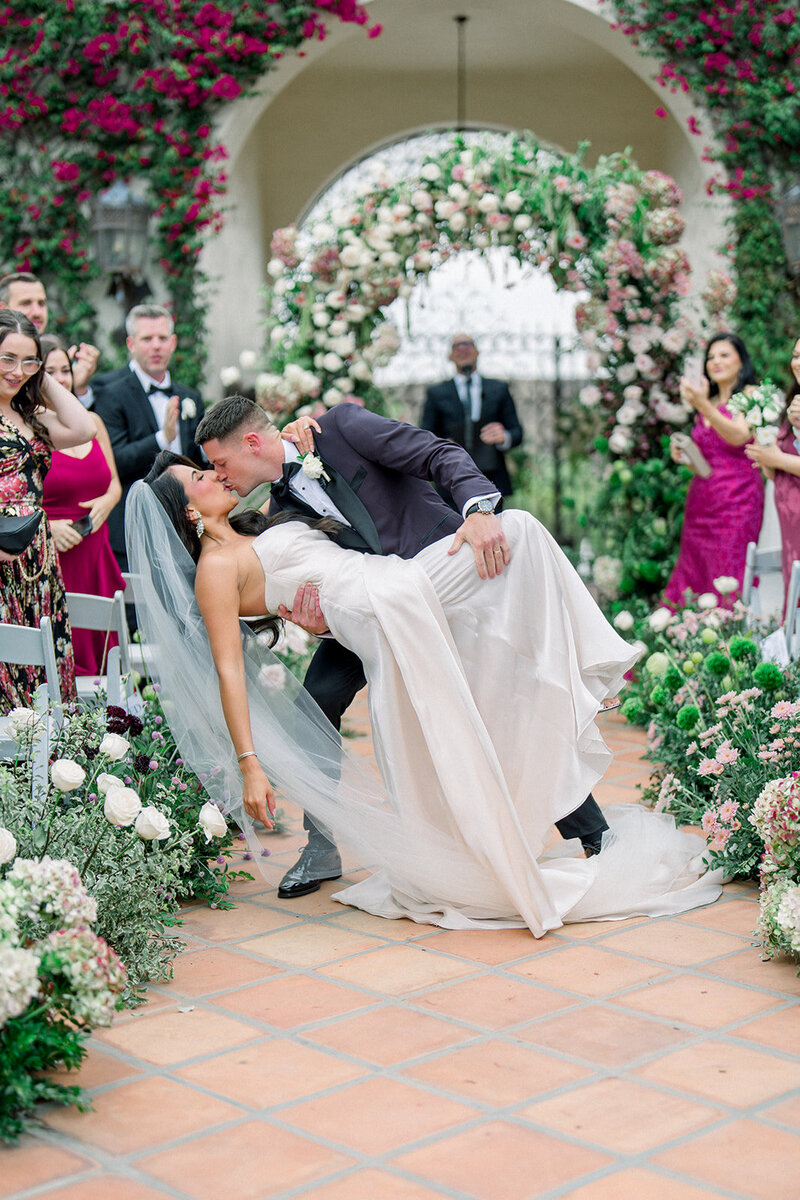 Groom dips and kisses bride in front of lush floral arch during luxury wedding ceremony in Southern California