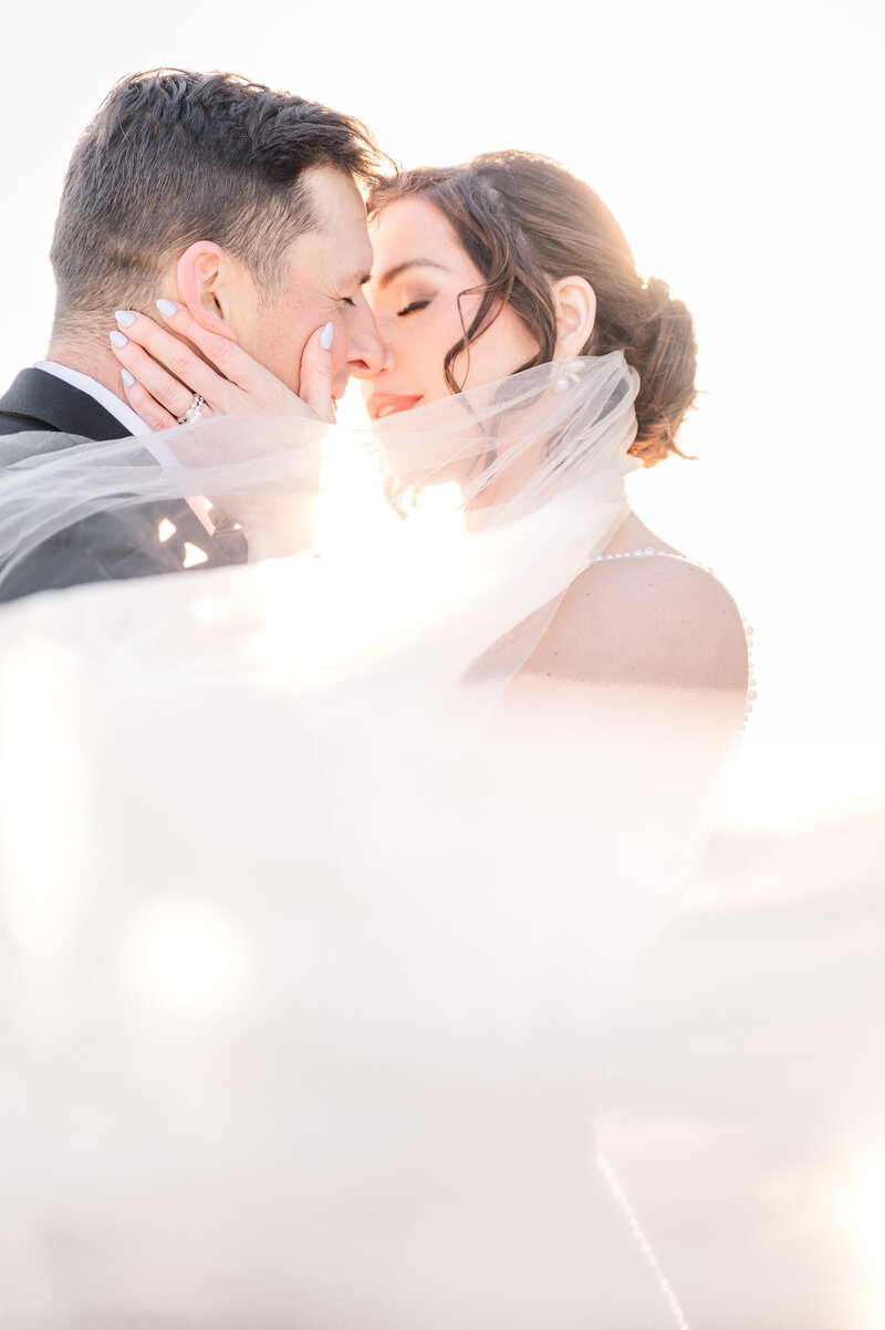 A laughing brunette bride with her husband as he kisses her cheek with her veil sweeping around them in June by JoLynn Photography