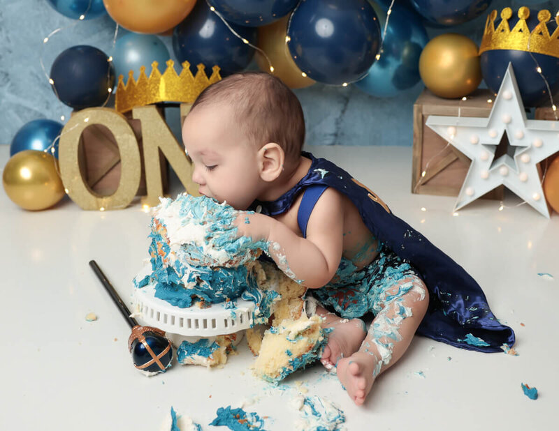 A joyful boy with icing on his face, surrounded by blue and gold balloons, engaged in a playful cake smash during a photography session in a Rochester, NY studio.