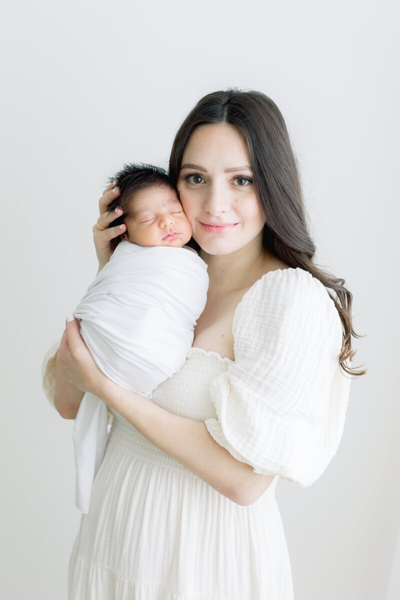Mom in white gown holding swaddled baby by Main Line Newborn Photographer