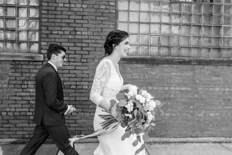 Groom wears sunglasses while walking in front of an urban brick building with his bride
