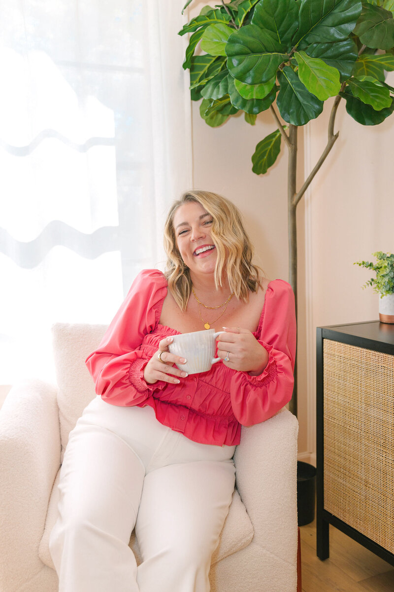 Mollie Mason in a pink top sitting in a white chair smiling and holding a white mug
