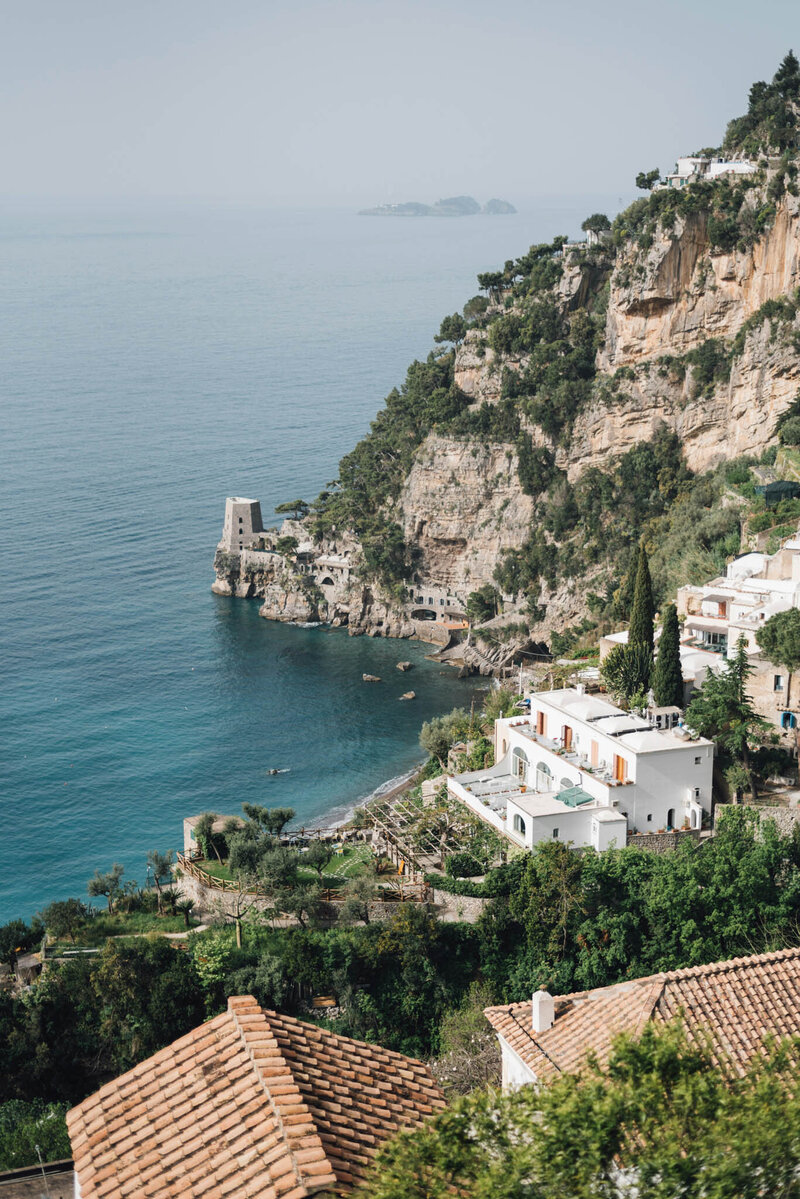 Scenic view of a coastal cliff with lush greenery and white houses overlooking a calm sea. The foreground has terracotta rooftops, and a medieval tower is visible on the rocky shore. The horizon is hazy under a clear sky.