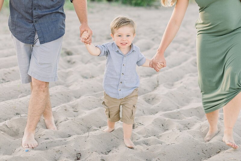 A toddler boy in a blue shirt and tan shorts is smiling at the camera as he holds his mom and dad's hands