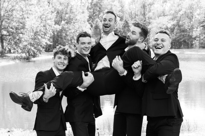 This is a fun and candid photo of a groom and his groomsmen being lifted in the air Photographed by GreenPoint Photography. The photo is in black and white, creating a classic and timeless look. The photo also shows the beautiful scenery of the lake and the trees in the background.