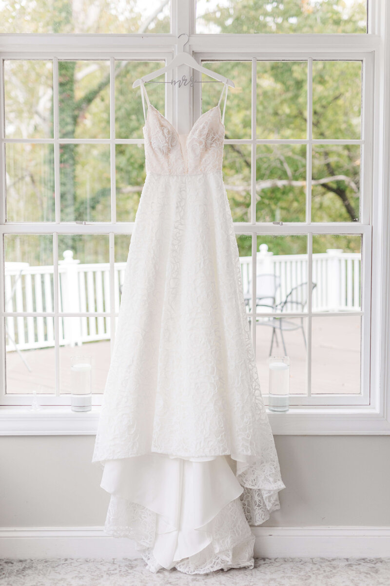 A brides dress hangs in the window of the wedding venue. It's a beautiful day outside and you can see the green trees.
