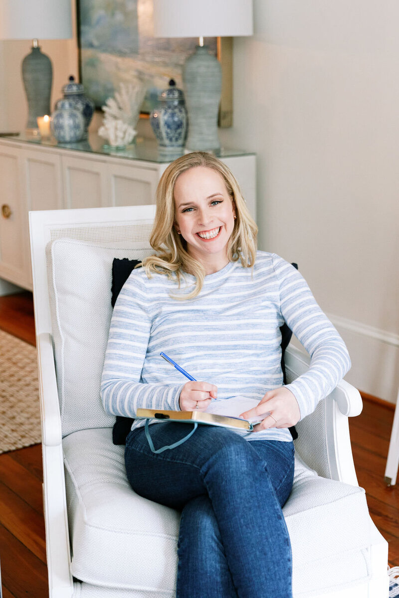 Sarah, founder of Digital Grace Design, smiling for the camera while sitting in ivory arm chair in living room wearing a blue and white striped sweater and jeans