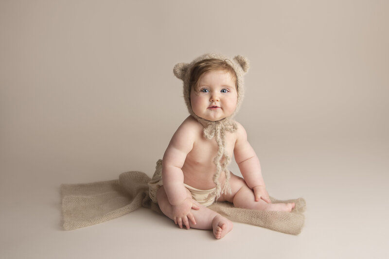 Smiling six month old baby girl wearing  a teddy bear knitted bonnet, sitting on a simple beige knitted wrap on a beige background.
