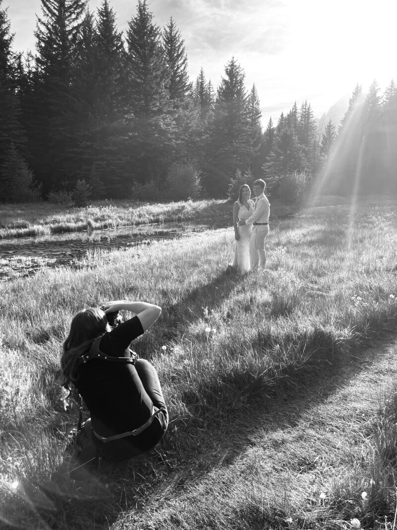 jackson hole photographers photographs bride adn groom embracing each other in a field in the grand tetons next to the Snake River  with the mountains in the distance as the photographer crouches and captures the moment