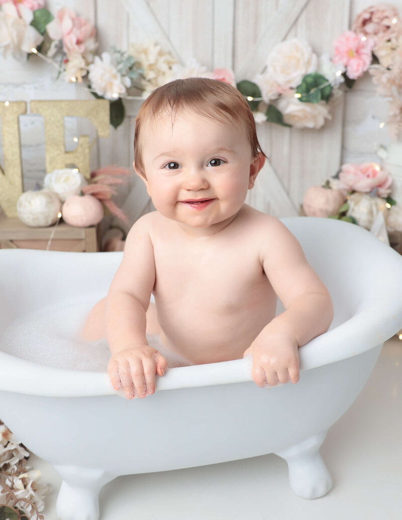 A joyful one-year-old girl splashes water during a floral cake smash photography session in a Rochester NY studio, surrounded by floral and pumpkin decorations.