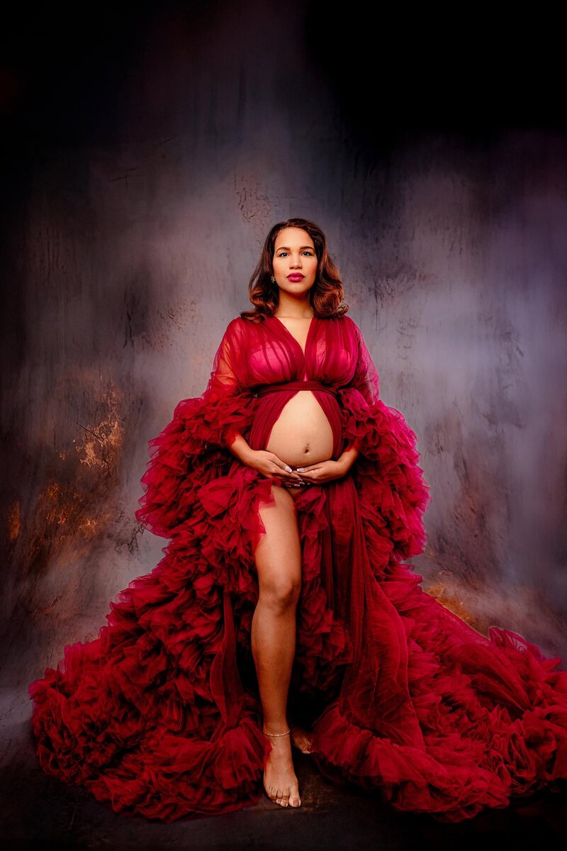 Expectant mom poses in Charlotte Studio in a red dress with both hands on her belly and her leg extended with her belly showing