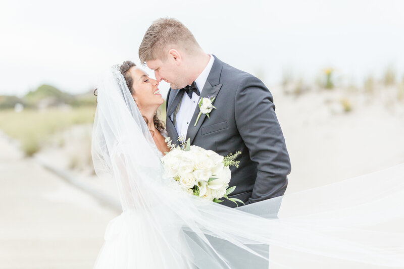 Bride and groom are photographed for an intimate couples portrait. They are facing each other with their noses touching and smiling. The bride's veil is blowing in front of them. Photo captured at Rhode Island's Dunes Club in Narragansett by best New England Wedding Photographer Lia Rose Weddings