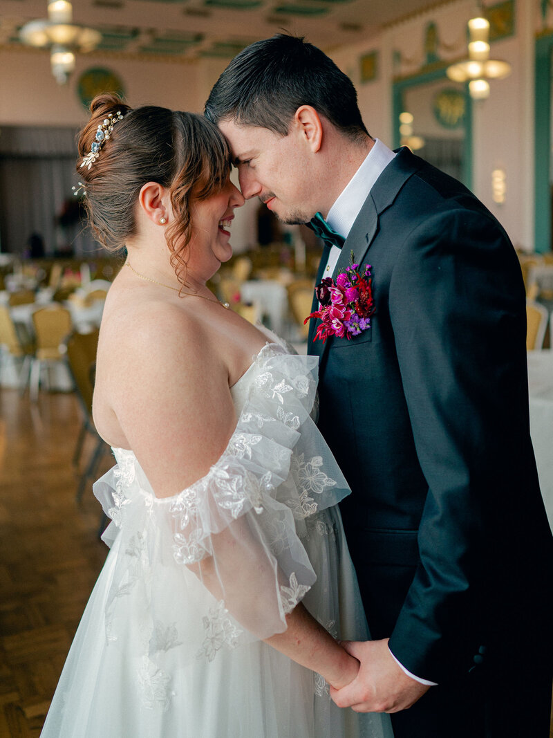 Bride and groom hold hands, foreheads together in reception space.