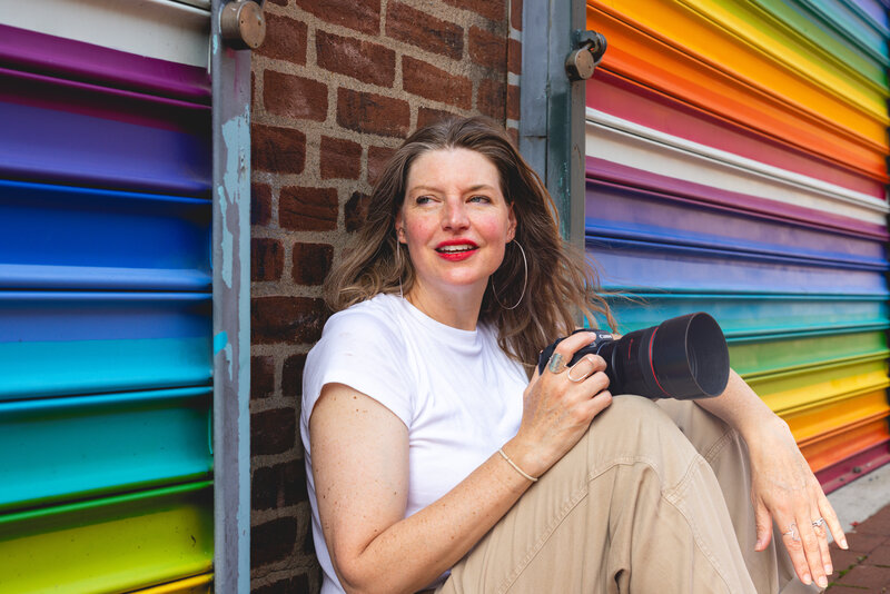 photo of woman against brick wall next to rainbow wall holding a camera