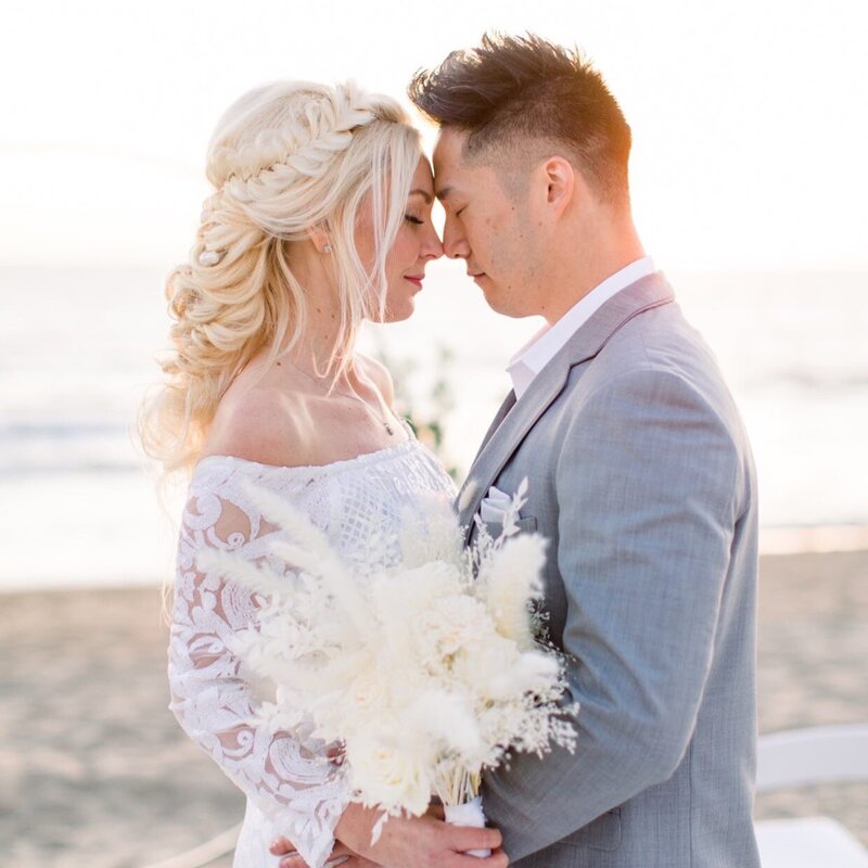 A bride and groom with foreheads touching at the beach with  the sun  coming in from the right side.