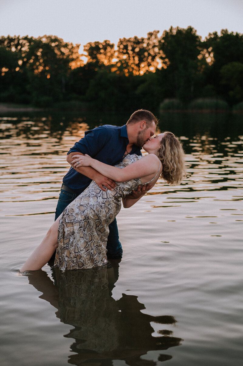 Engaged man dipping fiance and kissing her in a body of water at sunset