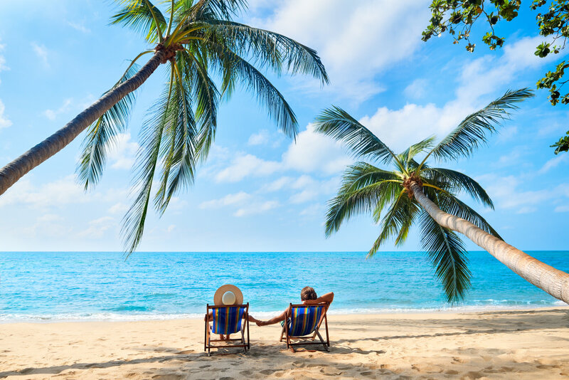 Two people relax on recliners on a sandy beach, facing a calm blue ocean. Palm trees arch over them, providing shade. The sky is clear with a few clouds, creating a serene tropical setting.