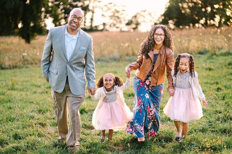 A happy family walks hand in hand in a field in Westchester; a man in a suit and a woman in a leather jacket and floral dress each hold hands with one of their two delighted daughters in pink skirts.