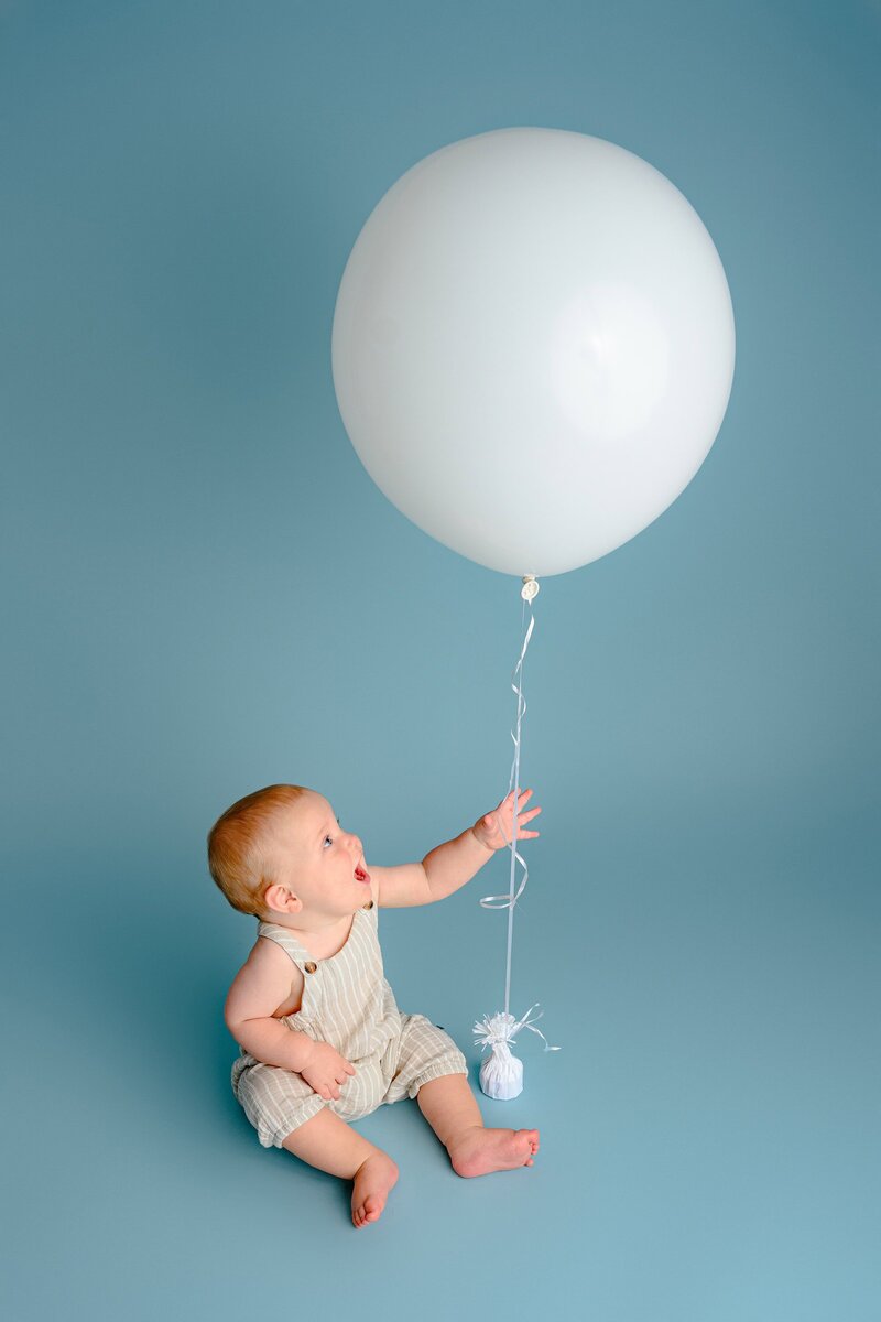 little boy sits in brown overalls holding large white balloon as he looks ip at it