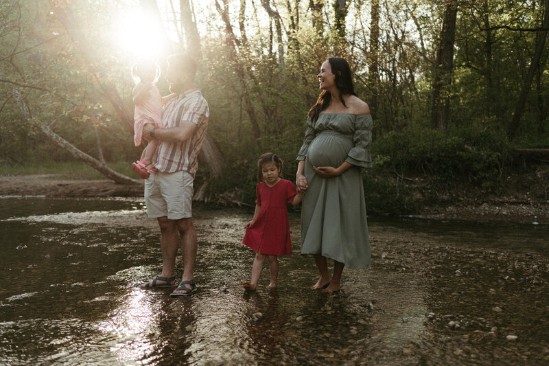A pregnant mom holding a toddler girls hand and a dad holding a toddler girl stand barefoot in a creek laughing at sunset
