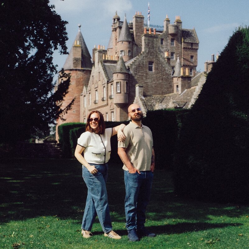 A husband and wife photography duo stand together on the lawn of a historic castle, smiling in the sun with the grand architecture behind them.
