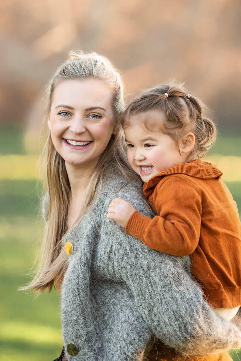 Mom in grey cardigan with daughter on her back both laughing in the park
