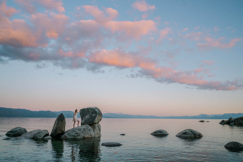 senior girl grad portrait  in a white dress on the rocks boulders in lake Tahoe at sunset pink skies